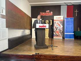 Pokaż zdjęcie: Za drewnianą mównicą stoi mężczyzna w marynarce krawacie i białej koszuli. Mówi do mikrofonu zamontowanego na stojaku.