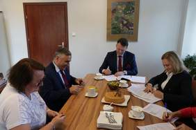 Pokaż zdjęcie: Przy dużym konferencyjnym stole siedzą dwie kobiety i dwóch mężczyzn. Wszyscy się uśmiechają . panowie ubrani w granatowe marynarki, białe koszule i krawaty. Po prawej stronie kobieta w blond włosach, również w granatowej marynarce i białej k