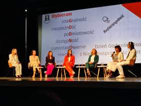 Pokaż zdjęcie: Panel dyskusyjny w którym bierze udział siedem kobiet, siedzących na krzesełkach ustawionych na scenie. W tle slajd z prezentacji z hasłami: integralność, niezależność, podmiotowość, dostępność, samostanowienie, równość.