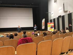 Przed zgromadzoną publicznością stoi dwóch mężczyzn i jeden z nich przema przez mikrofon