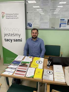 Pokaż zdjęcie: Mężczyzna siedzi przy stole, na którym znajdują się ulotki. Za nim baner promujący PFRON