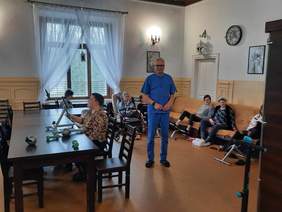 Pokaż zdjęcie: W sali znajdują sie osoby z niepełnosprawnościami, które ćwiczą na różnych urządzeniach. Na środku pokoju stoi pan rehabilitant, który nadzoruje wykonywane ćwiczenia. 