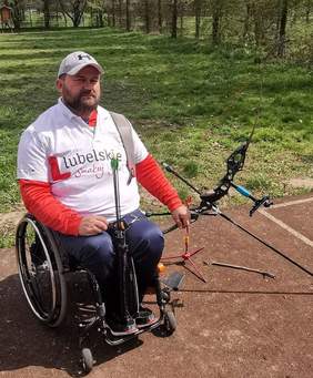 Pokaż zdjęcie: Mężczyzna na wózku inwalidzkim ,lewa ręka oparta na łuku. Między kolonami trzyma strzały 