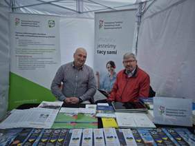 Pokaż zdjęcie: Pod namiotem znajduje się stoisko konsultacyjno-informacyjne Łódzkiego Oddziału PFRON. Przy stoisku siedzi na krzesłach dwóch mężczyzn, którzy się uśmiechają. 