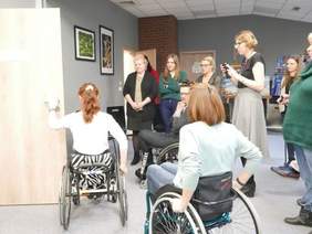 Pokaż zdjęcie: Trzy osoby poruszające się na wózkach inwalidzkich w holu. Jedna z nich otwiera drewniane drzwi. Obok stoją pozostali uczestnicy szkolenia.