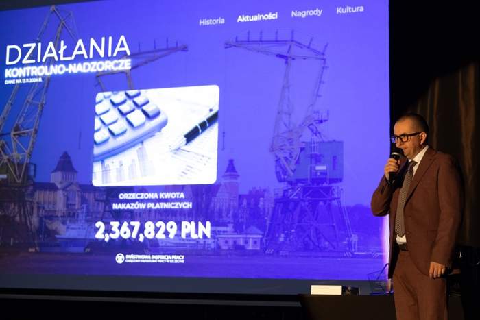 Pokaż zdjęcie: Mężczyzna w brązowym garniturze stoi na scenie przed dużym ekranem, na którym wyświetlony jest ten sam tekst: "RAZEM DLA BEZPIECZEŃSTWA - PREWENCJA 2024". Wygłasza przemówienie.