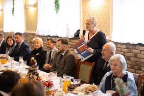 Pokaż zdjęcie: Na zdjęciu uroczyste spotkanie przy długim stole zastawionym jedzeniem i napojami. Uczestnikami spotkania są seniorzy oraz osoby w średnim wieku, wszyscy elegancko ubrani. Kobieta stojąca przy stole trzyma mikrofon i dokumenty, przemawia do zgromadzonych.