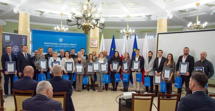 Pokaż zdjęcie: W sali kolunowej do pamiątkowego zdjęcia pozują 23 osoby, które trzymają w ręku pamiątkowe dyplomy.