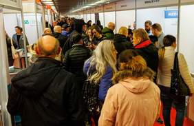 Pokaż zdjęcie: Zatłoczony korytarz podczas Targów Pracy w Białymstoku. Uczestnicy, ubrani w kurtki, przemieszczają się między stoiskami firm i instytucji. W tle widoczne są stoiska z nazwami firm oraz przedstawiciele pracodawców prowadzący rozmowy z odwiedzającymi.