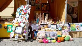 Pokaż zdjęcie: Na Rynku Starego Miasta pod namiotem na stoliku wystawione sąa kolorowe prace uczestników warsztatu terapii zajęciowej. Są to: maskotki, rzeźby, biżuteria.