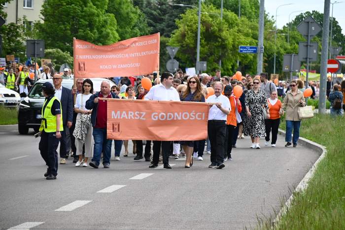 Pokaż zdjęcie: Marsz Godności w Zamościu – uczestnicy idą ulicą z pomarańczowymi balonami i transparentem „Marsz Godności”; na czele grupa niesie baner stowarzyszenia, wydarzenie zabezpiecza policja.