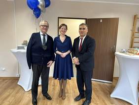 Pokaż zdjęcie: Kobieta w eleganckiej sukience. Po bokach stoi dwóch mężczyzn w białych koszulach i krawatach, Jeden ma na sobie garnitur drugi granatowy sweter. Za nimi kolorowe balony, obok stół z kateringiem.