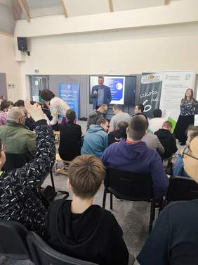 Pokaż zdjęcie: Zdjęcie z tyłu sali w kierunku osób prowadzących konkurs. Młodzież siedzi na ławeczkach, bierze aktywny udział w spotkaniu.