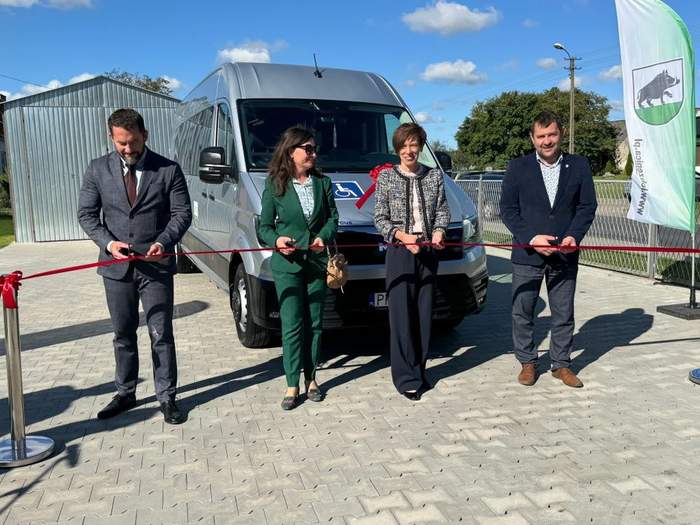 Pokaż zdjęcie: Dwie kobiety oraz dwóch mężczyzn przecina czerwoną wstęgę. Z tyłu znajduje się blaszany garaż oraz srebrny bus z naklejką informującą, że pojazd przystosowany jest do przewozu osób z niepełnosprawnościami.