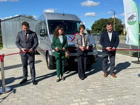 Pokaż zdjęcie: Dwie kobiety oraz dwóch mężczyzn przecina czerwoną wstęgę. Z tyłu znajduje się blaszany garaż oraz srebrny bus z naklejką informującą, że pojazd przystosowany jest do przewozu osób z niepełnosprawnościami.