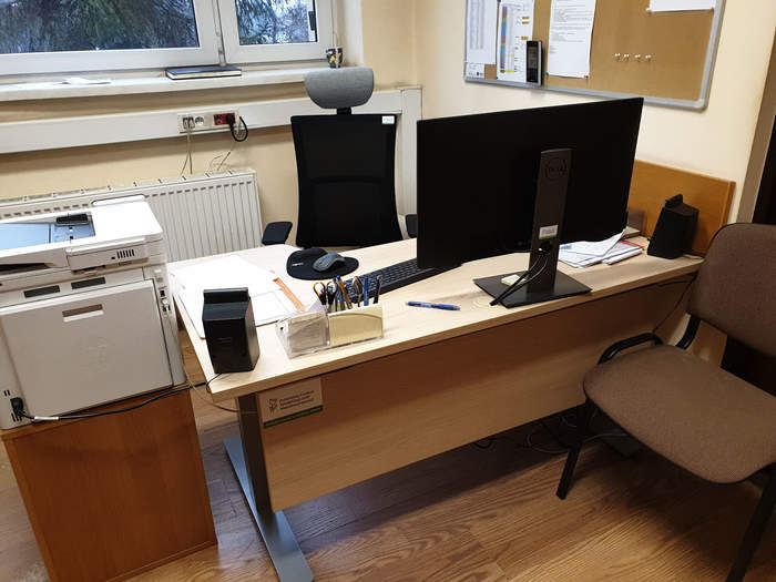 Pokaż zdjęcie: fragment pomieszczenia z biurkiem na nim stoi duży monitor, klawiatura, rozłożone papiery obok na kontenerze stoi wielofunkcyjne urządzenie przed biurkiem stoi brązowe krzesło a za biurkiem czarny fotel na biurku naklejone jest logo PFRON
