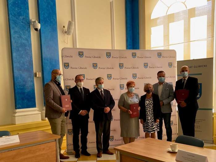 Pokaż zdjęcie: Fragment dużej sali siedem osób stoi na tle ścianki z herbami i napisami powiat gliwicki 3 osoby trzymają teczki 