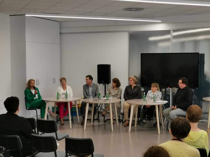 Pokaż zdjęcie: Przy trzech okrągłych stołach siedzi siedem osób. Jedna z kobiet siedzi na wózku inwalidzkich. Pierwsza kobieta od lewej trzyma w ręku mikrofon, pozostałe skierowały wzrok na nią.  