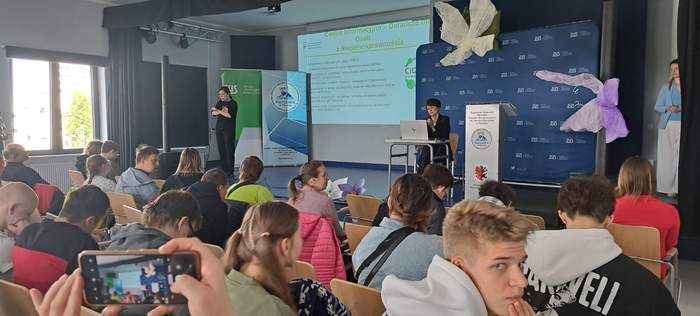 Uczestnicy wydarzenia siedzą na sali i słuchają prelegentki. W tle widać ekran z prezentacją, a po lewej stronie widać tłumaczkę języka migowego.