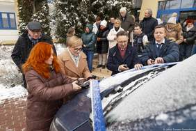 Pokaż zdjęcie: Grupa osób stojących przy nowym mikrobusie przecina wstęgę podczas uroczystości przekazania pojazdu przed budynkiem Domu Pomocy Społecznej w zimowej scenerii.