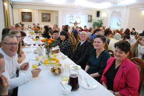 Uśmiechnięci uczestnicy wydarzenia siedzą przy zastawionych stołach, świętują