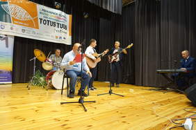 Występ sceniczny 5 mężczyzn, grają na gitarach, perkusji, instrumencie klawiszowym, jeden z tekstem w rękach