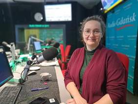 Pokaż zdjęcie: W studio radiowym przy mikrofonie siedzi dziewczyna w okularach. Ma na sobie ciemny golf i sweterek. Uśmiecha się. W tle logo Radia Gdańsk.