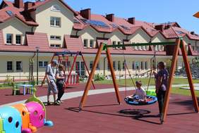 Pokaż zdjęcie: Plac zabaw gdzie jeden chłopiec huśta się na huśtawce z asekuracją pani a drugi trzymany za rękę przez druga panią spaceruje