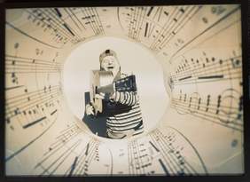 Pokaż zdjęcie: Mężczyzna z zespołem downa trzyma gitarę z której celuje jak z karabinu w stronę otaczającego go rysunku pięciolinii z nutami.