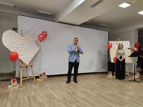 Pokaż zdjęcie: Mężczyzna przemawia do zgromadzonych, trzymając mikrofon. Po bokach sceny stoją dekoracje z czerwonymi balonami, sercem z nazwiskami oraz tablica z napisem „Otwarte Serca 2026”. Obok stoi kobieta przy mikrofonie.