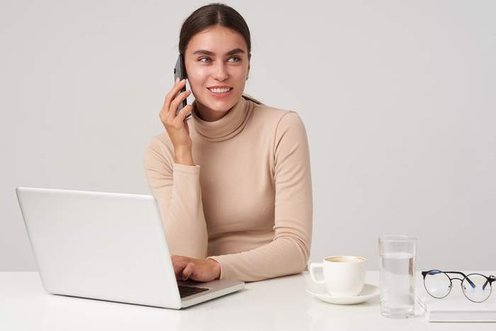 Pokaż zdjęcie: Kobieta siedzi przy laptopie. Trzyma słuchawkę .Przed nią otwarta książka 