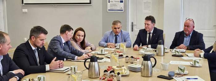 Pokaż zdjęcie: Spotkanie przy dużym stole konferencyjnym; kilkanaście osób w garniturach rozmawia, notatniki i dokumenty obok filiżanek kawy oraz dzbanków na środku; jasna sala z ekranem projekcyjnym i drzwiami w tle.