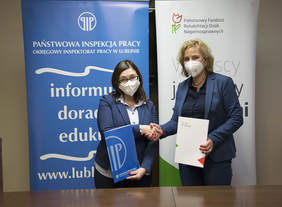 Pokaż zdjęcie: dwie kobiety w niebieskich marynarkach i maseczkach stoją przy brązowym stole, wymieniają uściski, w tle loga PIP oraz PFRON