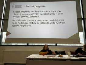 Pokaż zdjęcie: Kobieta siedzącą za stołem prezydialnym, na ekranie wyświetla się prezentacja