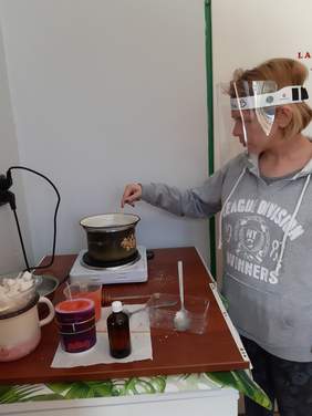 Pokaż zdjęcie: Dziewczyna stoi przy stoliku miesza patyczkim ciecz w garnku stojącym na elektrycznej płycie obok stoją pełne i puste pojemniki, butelka, garnek