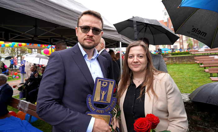 Zbliżenie na dwoje uśmiechniętych ludzi stojących obok siebie w deszczowy dzień. Mężczyzna w okularach przeciwsłonecznych i granatowej marynarce trzyma pamiątkową statuetkę z napisem „Otwórzmy przed nimi życie”. Kobieta obok niego ma na sobie jasną marynarkę i trzyma czerwone róże. W tle widać kolorowe balony, parasole i inne osoby uczestniczące w wydarzeniu plenerowym.