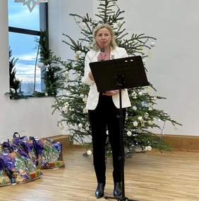 Pokaż zdjęcie: Przy mikrofonie stoi kobieta ubrana w białą marynarkę i czarne spodnie, składa życzenia.