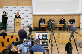 Pokaż zdjęcie: Przemawiająca przy mównicy kobieta w sali wykładowej. Za nią wyświetlona prezentacja multimedialna i 4 siedzących mężczyzn. Wydarzenie nagrywane i transmitowane przez zgromadzonych techników. 