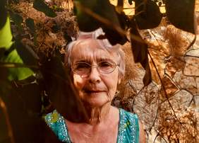 Pokaż zdjęcie: Zdjęcie babci Zosi, uśmiechniętej starszej kobiety w okularach, wychylającej się zza liści drzewa