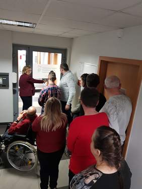 Pokaż zdjęcie: Grupa dziewięciu osób stoi w wąskim korytarzu budynku. Uczestnicy są zwróceni tyłem do obiektywu, słuchając kobiety, która wskazuje ręką na piktogramy przy drzwiach. Na pierwszym planie po lewej stronie widoczna jest osoba na wózku. Jasne ściany korytarza kontrastują z brązowymi drzwiami, a w tle widać przeszklone drzwi, przez które wpada światło.