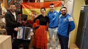 Pokaż zdjęcie: Mężczyzna z opaską na oczach i w hiszpańskim czarnym kapeluszu ubrany na czarno, z czerwoną much siedzi i trzyma akordeon. Obok dziewczyna w czerwonej sukni i dwóch mężczyzn w niebieskich bluzach. 