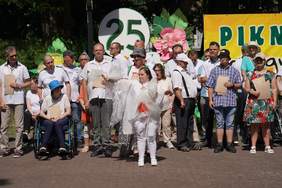 Pokaż zdjęcie: Grupowe zdjęcie uczestników plenerowego pikniku jubileuszowego. Kilkanaście osób w letnich strojach stoi i siedzi w rzędach, w tym jedna osoba na wózku. W tle widać dekoracje: białe koło z zieloną liczbą „25”, duże różowe kwiaty, fragment żółtego jubileuszowego banneru z napisem oraz gęste drzewa.
