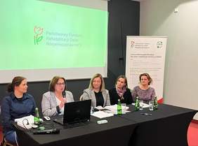 Pracownice Oddziału Kujawsko-Pomorskiego PFRON siedzą przy stole. Jedna z nich prowadzi spotkanie. Z tyłu widoczny jest ekran i baner z logo PFRON