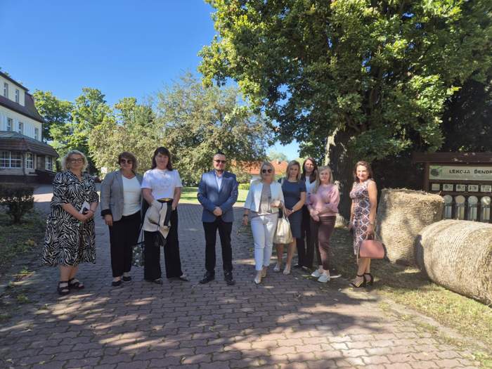 Pokaż zdjęcie: Grupa kobiet i jeden mężczyzna pozuje do zdjęcia, stojąc obok siebie na terenie posesji Domu Pomocy Społecznej, a w tle ładna pogoda i świecące słońce wraz z otaczającą zielenią