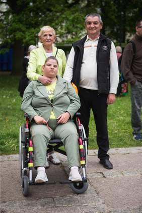 Pokaż zdjęcie: Na zdjęciu pan z panią przed nimi wózek inwalidzki na którym siedzi dziewczyna. Rodzice się uśmiechają w tle drzewa i uczestnicy wydarzenia.