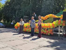 Pokaż zdjęcie: Trzy osoby stoją przy scenie ozdobionej kolorowymi balonami podczas plenerowego wydarzenia. Mężczyzna przemawia do mikrofonu w słoneczny dzień.