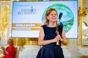 Pokaż zdjęcie: Kobieta w granatowej sukni trzyma statuetkę i uśmiecha się podczas gali. W tle ekran z napisem „Człowiek bez Barier” oraz nazwiskiem Izabela Dłużyk, złocona sala i siedząca osoba bijąca brawo.  