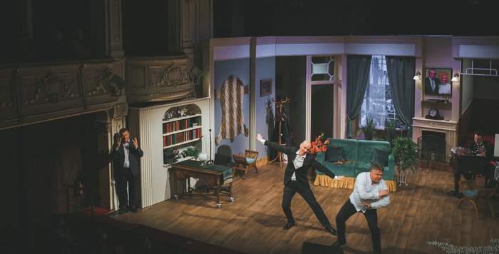 Pokaż zdjęcie: Na scenie teatru tańczy dwóch mężczyzn za nimi scenografia przedstawiająca umeblowany pokój. Z boku stoi tłumacz języka migowego.