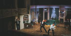 Pokaż zdjęcie: Na scenie teatru tańczy dwóch mężczyzn za nimi scenografia przedstawiająca umeblowany pokój. Z boku stoi tłumacz języka migowego.