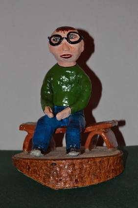 Pokaż zdjęcie: Rzeźba człowieka siedzącego na ławeczce. Postać ma okulary, niebieskie spodnie, zielony sweter i tenisówki